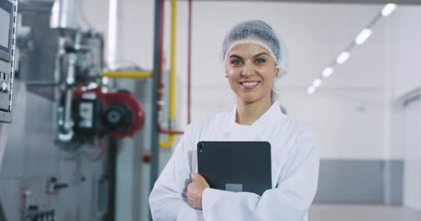 大きな製造業工場で電子タブレットを持ちながら、カメラをまっすぐ見つめ、大きく微笑む成熟した女性魅力的なエンジニアのクローズアップポートレート — ストック動画