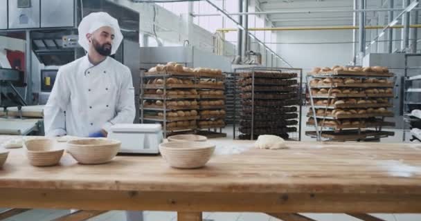 Panadero carismático bailando con barba en un elegante uniforme en la industria de panadería grande mientras escucha música de auriculares inalámbricos — Vídeos de Stock