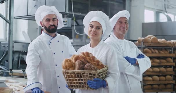 Snygg kvinna med ett stort leende Baker och hennes kollegor bagare i en vit Uniform stående bredvid den stora industri maskinen och hålla en korg med ekologiskt bröd — Stockvideo