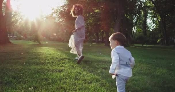Молодая семья в стильных костюмах, играющая посреди парка, улыбается двум маленьким детям, бегущим за родителями — стоковое видео