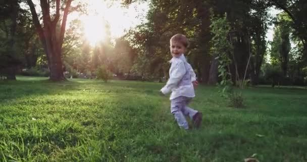 Маленький смішний хлопчик на заході сонця в парку через зелену траву в милому костюмі — стокове відео