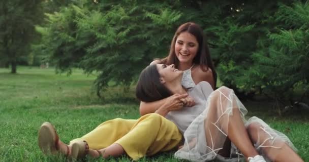 Привлекательные две дамы в парке наслаждаются временем вместе сидя на траве и болтая улыбаясь большой рядом с камерой — стоковое видео