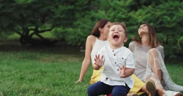 Very cute two years boy make funny faces in front of the camera while on the background two ladies smiling large and enjoying the time spending in the park — Stock Video