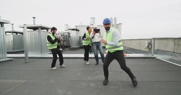 Egy csodálható építési ember élvezi az idejét a barátaival egy építési tető tetején, míg egy férfi, ha egy fehér inget filmek őket a tabletta — Stock videók