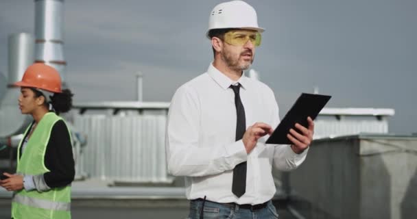 A good looking old chubby construction man wears a white helmet and shirt with a black tie, he taps on his tablet while a pretty young woman behind him talks on her transceiver — Stock Video