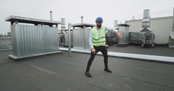 En el sitio de construcción en la parte superior del edificio carismático hombre ingeniero o arquitecto disfrutando del momento bailando excitado con un casco de seguridad y una ración en la mano — Vídeo de stock