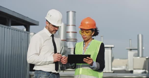 Viejo ingeniero y su mujer de asistencia étnica africana analizando el plan de construcción en la azotea del edificio hablando por ración con el capataz, que llevaba equipo de seguridad — Vídeos de Stock