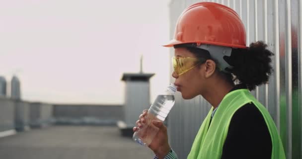 美丽的女工程师带着头盔和安全眼镜口渴在建筑工地的屋顶上喝水 — 图库视频影像