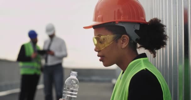 Удивительная женщина инженер очень пить питьевую воду из пластиковой бутылки на крыше здания на строительном фоне другие работники имеют обсуждение — стоковое видео
