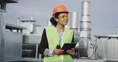 Turuncu kask ve sarı güvenlik gözlükleri giyen inşaat alanının çatısında bir tablet ve istihkak tutan şantiyede Çekici kadın Afrikalı etnik genç iş kadını