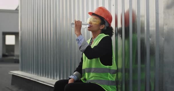 Харизматичная африканская женщина-инженер улыбается, выпивая немного воды на крыше на фоне строительства ее коллеги анализируют план строительства — стоковое видео