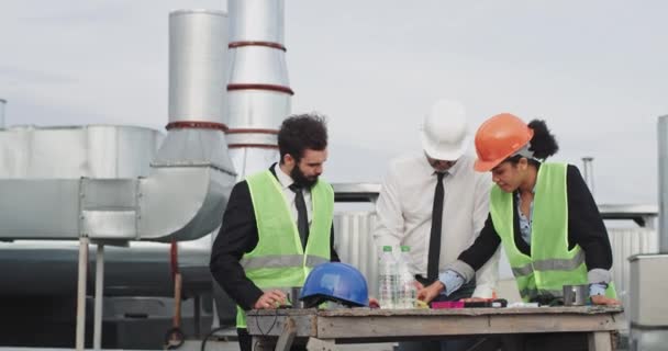 Auf dem Dach der Baustelle analysieren zwei Ingenieure und ein Architekt multiethnisch auf dem Tisch den Bauplan des Gebäudes, in dem sie Schutzhelme tragen — Stockvideo