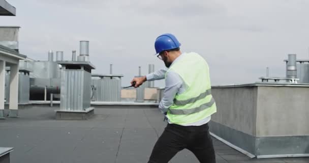 Забавный красивый архитектор на крыше здания танцует и чувствует себя очень взволнованным в шлеме безопасности и деловой одежде — стоковое видео