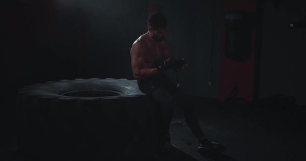 In the dark cross fitness class athletic guy after his intense workout training take off the professional bandages while sitting on the large tire — Stock Video