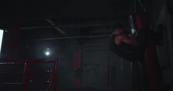 In een donkere boksklas cross fit concept atletische jonge man doet buikspieroefeningen terwijl het vasthouden met de benen van de bokszak — Stockvideo