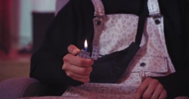 Kanepede oturan bir adam kameranın önünde çakmakla oynuyor, ateş yakıyor.