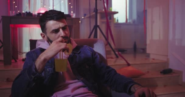 Охлаждающая атмосфера на домашней вечеринке, полной украшений, парень наслаждается временем, выпивая коктейль, сидя на диване — стоковое видео