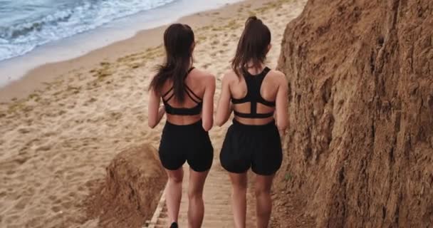 Las dos chicas en forma y deportivas corren a la playa y disfrutan de la compañía del otro mientras hacen deporte — Vídeos de Stock