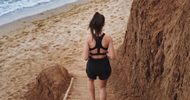 Een charismatisch jong dun meisje rent naar het strand en heeft zand op haar schoenen, ze draagt een korte broek en een sportbeha — Stockvideo