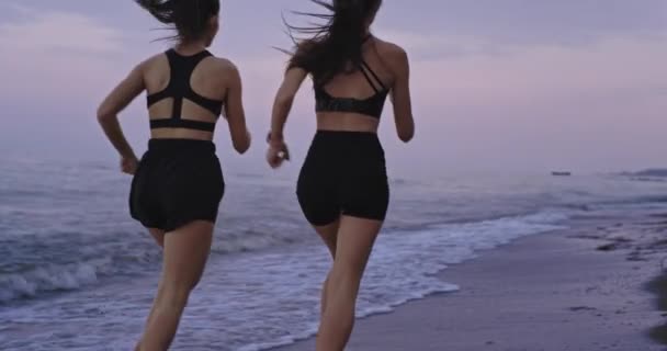 Atractivas dos jóvenes corriendo por la playa disfrutando del momento juntas usando ropa deportiva, escuchando música desde auriculares inalámbricos. despacio — Vídeo de stock