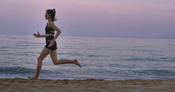 Sahilde koşan profesyonel bir bayan siyah bir spor giyim kuşamı giyerek sıkı koşuyor. — Stok video