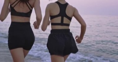 Sahilde birlikte koşan ve kablosuz kulaklıklardan müzik dinleyen spor giysili iki çekici bayan.