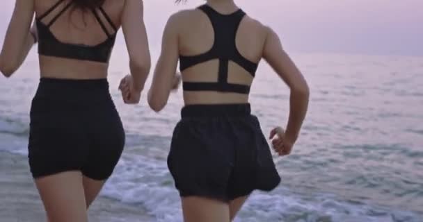 Zwei attraktive sportliche Damen in Sportbekleidung, die gemeinsam am Strand laufen und Musik aus den drahtlosen Kopfhörern hören — Stockvideo