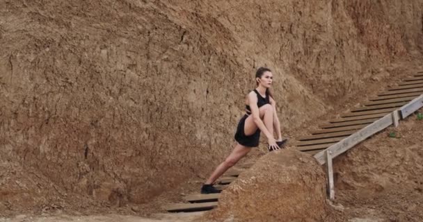 Twee dames in een zwarte Sportswear stretching benen op de houten stents naast het strand achtergrond grote rotsen — Stockvideo