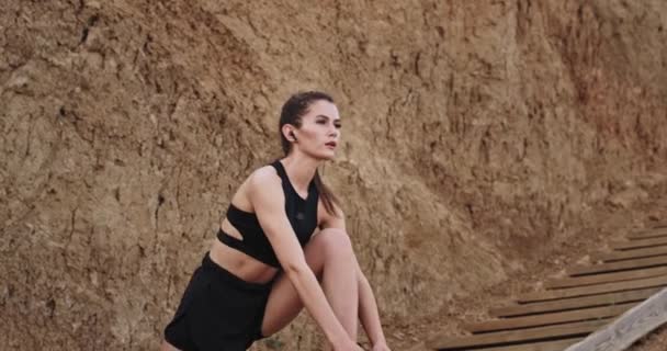 Schöne sportliche Dame hat ein Workout, das den Körper nach draußen in den hölzernen Stufen neben dem Meer streckt, hört sie Musik aus drahtlosen Kopfhörern — Stockvideo