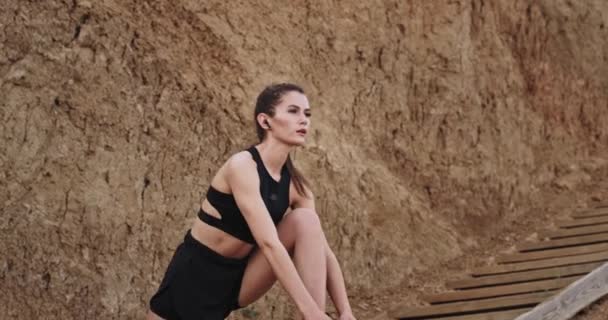 Belle dame sportive ont une séance d'entraînement étirant le corps à l'extérieur dans les marches en bois à côté de la mer, elle écoute de la musique à partir d'écouteurs sans fil — Video