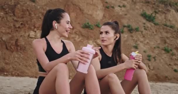 Charismatic young women have a conversation after the workout sitting down on the beach in front of the sea — ストック動画