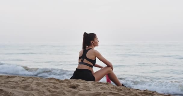 Gesundes Lifestylekonzept einer sportlichen Dame nach dem Training am Strand vor dem Meer, das durstige Wasser trinkend und das Leben genießend — Stockvideo