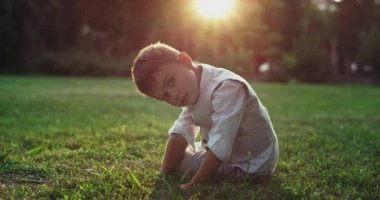 Very seriously face of a little boy 5 years old looking straight to the camera in the middle of the park falling down on the grass rolling start to smiling very cute