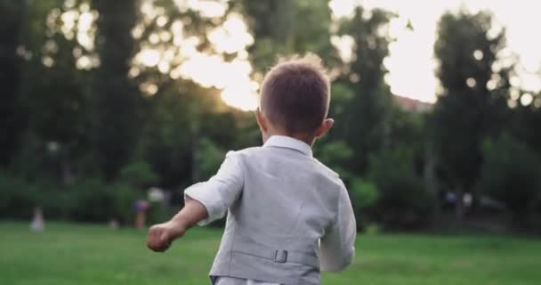 Маленький милий хлопчик у костюмі, що проходить через зелену траву посеред парку, насолоджуючись часом на природі — стокове відео