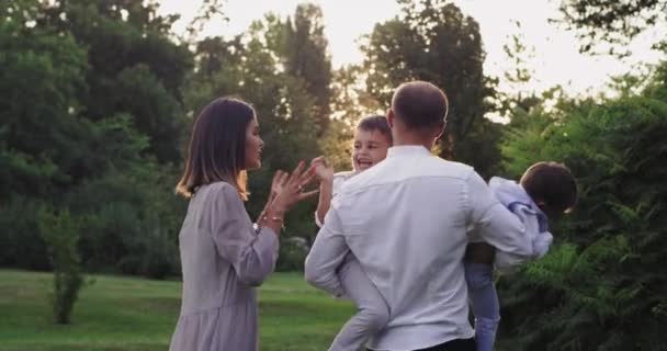 Молодая семья с двумя детьми, наслаждающаяся временем вместе посреди зеленого парка, широко улыбается и прыгает, обнимаясь, чувствуя себя счастливой перед камерой — стоковое видео