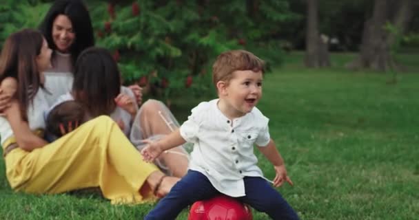 Забавный маленький мальчик сидит на красном фоне, его большая семья проводит прекрасное время в центре парка. — стоковое видео