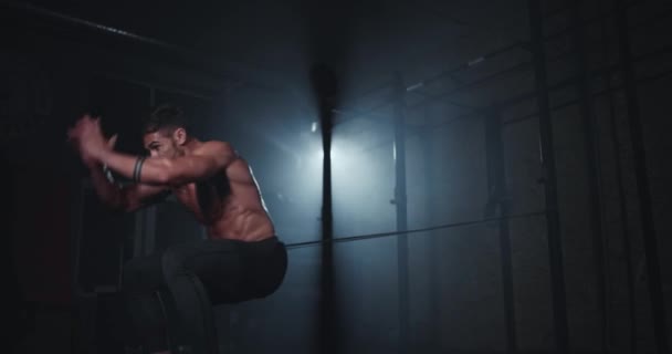Дуже жорсткий хрест вправи в темному тренажерному класі спортивний хлопець стрибає важко все пітніння, використовуючи еластичні смуги, щоб тримати його тіло — стокове відео