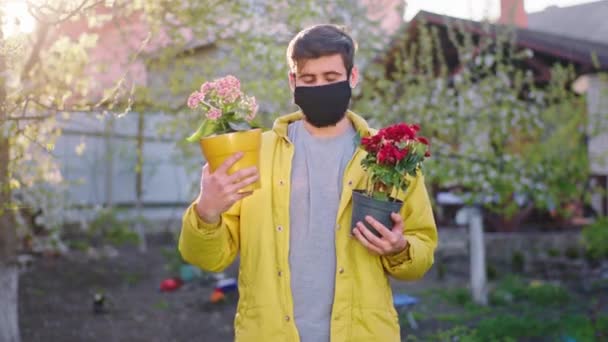 Killen i karantänen med en skyddsmask framför kameran håller två blomkrukor och tittar rakt mot kameran medan det är hemma i den pandemiska Coronavirus — Stockvideo