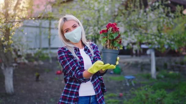 Snygg blond dam hemma i trädgården i karantän bär skyddsmask hon håller en blomkruka och tittar rakt in i kameran — Stockvideo
