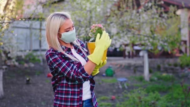 V zahradě v pandemické dámě s ochrannou maskou před kamerou drží květináč ona cítí radost starat se o květiny Covid -19 — Stock video