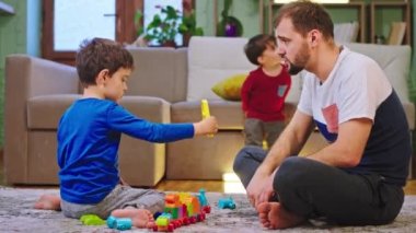 Çekici bir baba ve karizmatik oğlu oturma odasında yerde oturmuş arka planda televizyon seyreden diğer çocuklarla sohbet ediyorlar. ARRI Alexa Mini 'de çekilen.