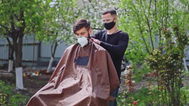 Ve velké zahradě dva přátelé pohlední kluci s ochrannou maskou mají účes čas jeden z nich stříhat vlasy do druhého v karanténě doma v zahradě Covid-19 — Stock video