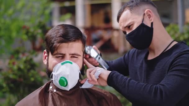 Professionele kapper man thuis in de tuin met een beschermend masker doet een kapsel voor een man die hij met behulp van de tondeuse — Stockvideo
