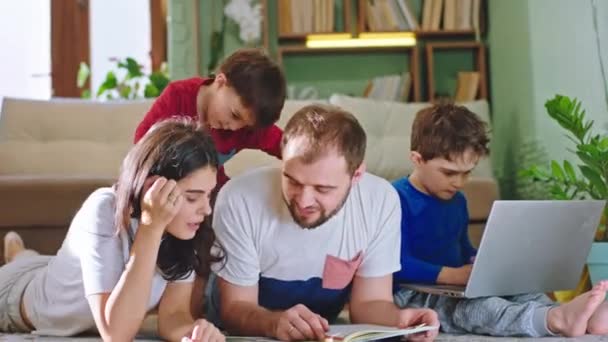 En la sala de estar carismático mamá y papá y dos niños lindos pasar un buen rato juntos mientras yacen en el suelo que preparan un libro de cuentos y otro niño grande jugando en la computadora portátil — Vídeo de stock