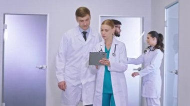 Büyük hastane koridorunda kameranın önünde. Ana doktor tablete bakıyor ve konuşuyor. Sarışın genç bayan doktorla bazı sonuçları analiz ediyor.