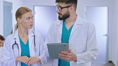 Çok ırklı iki doktorun önünde hastane koridorunda yürüyen iki kadın ve erkek meslektaşın dijital tablet kullanarak hastanın teşhisini tartışıyorlar.