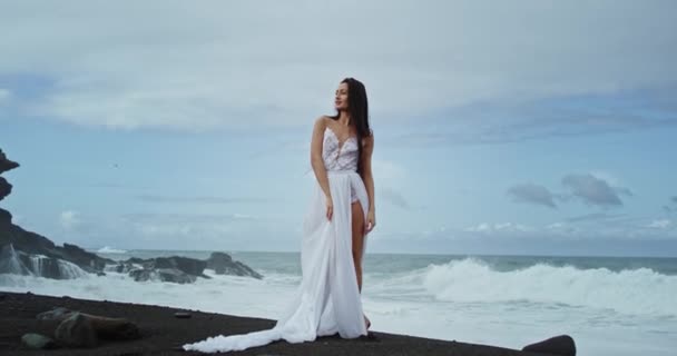 Mulher muito atraente em um vestido romântico branco ao lado do litoral ela olhando para o fundo da câmera paisagem incrível com rochas e ondas incríveis vista — Vídeo de Stock