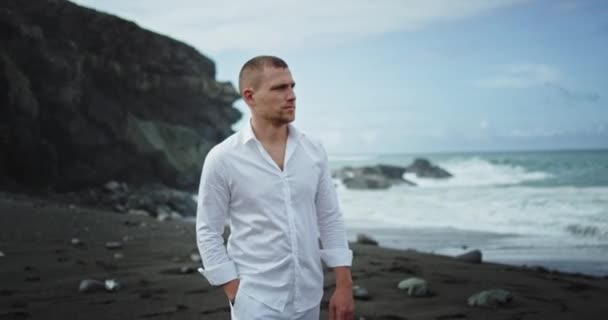 Homem bonito nas roupas brancas andando pela praia com areia preta ana paisagem incrível com grandes rochas e vista para o mar — Vídeo de Stock