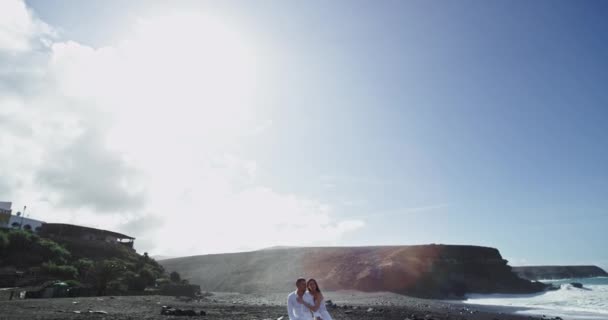 En un día soleado en medio de un paisaje increíble, una pareja de damas en un vestido largo y blanco y un hombre con una ropa blanca pasan el tiempo sentado en la roca junto al mar — Vídeos de Stock