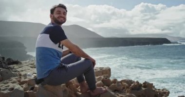 Büyük bir gülümsemesi olan çekici bir genç dağ ve denizin ortasında büyük dalgalarla harika bir manzaranın tadını çıkarıyor. 4k